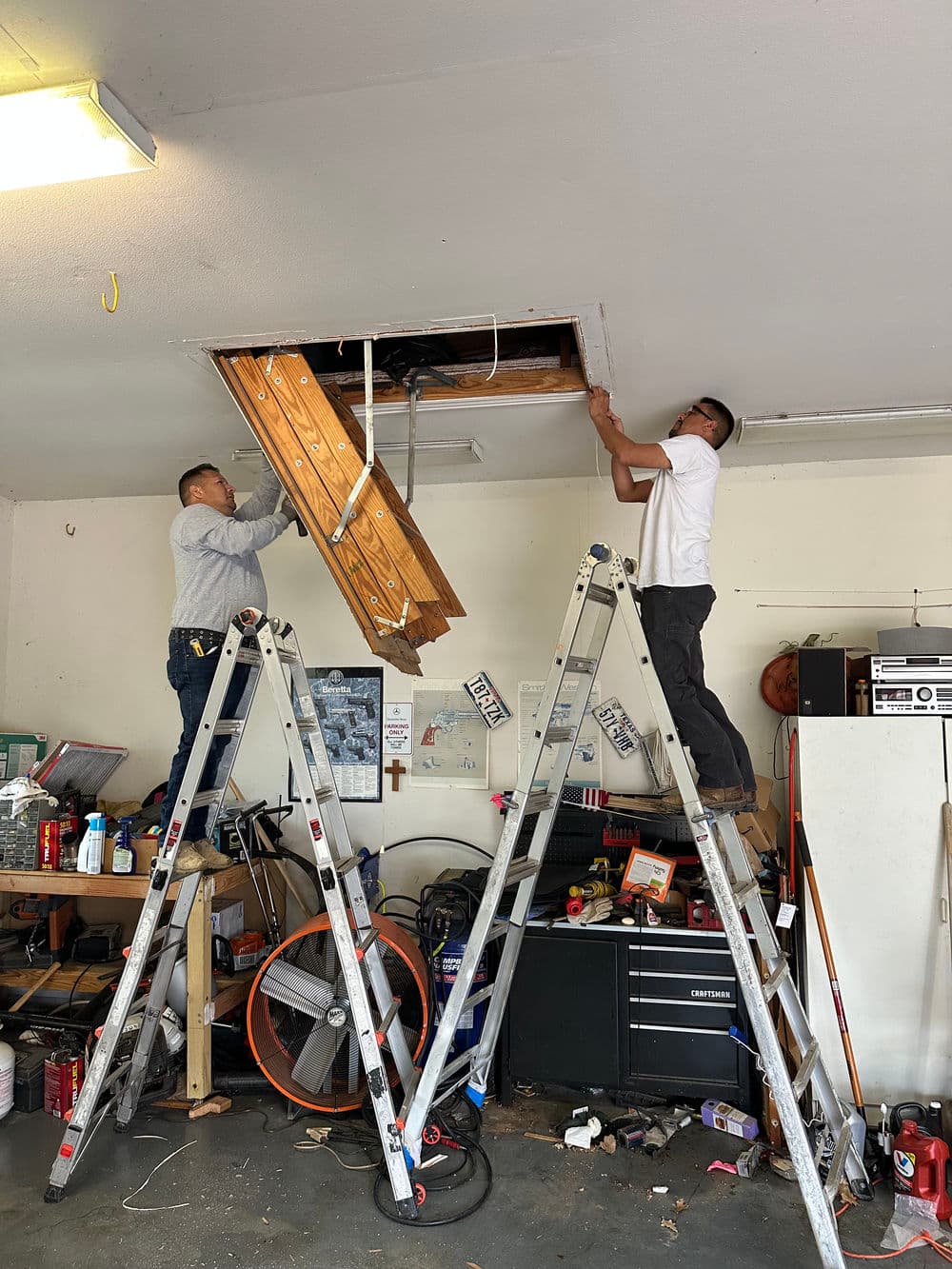 Two workers using ladders to repair a ceiling in a garage with tools and materials nearby.